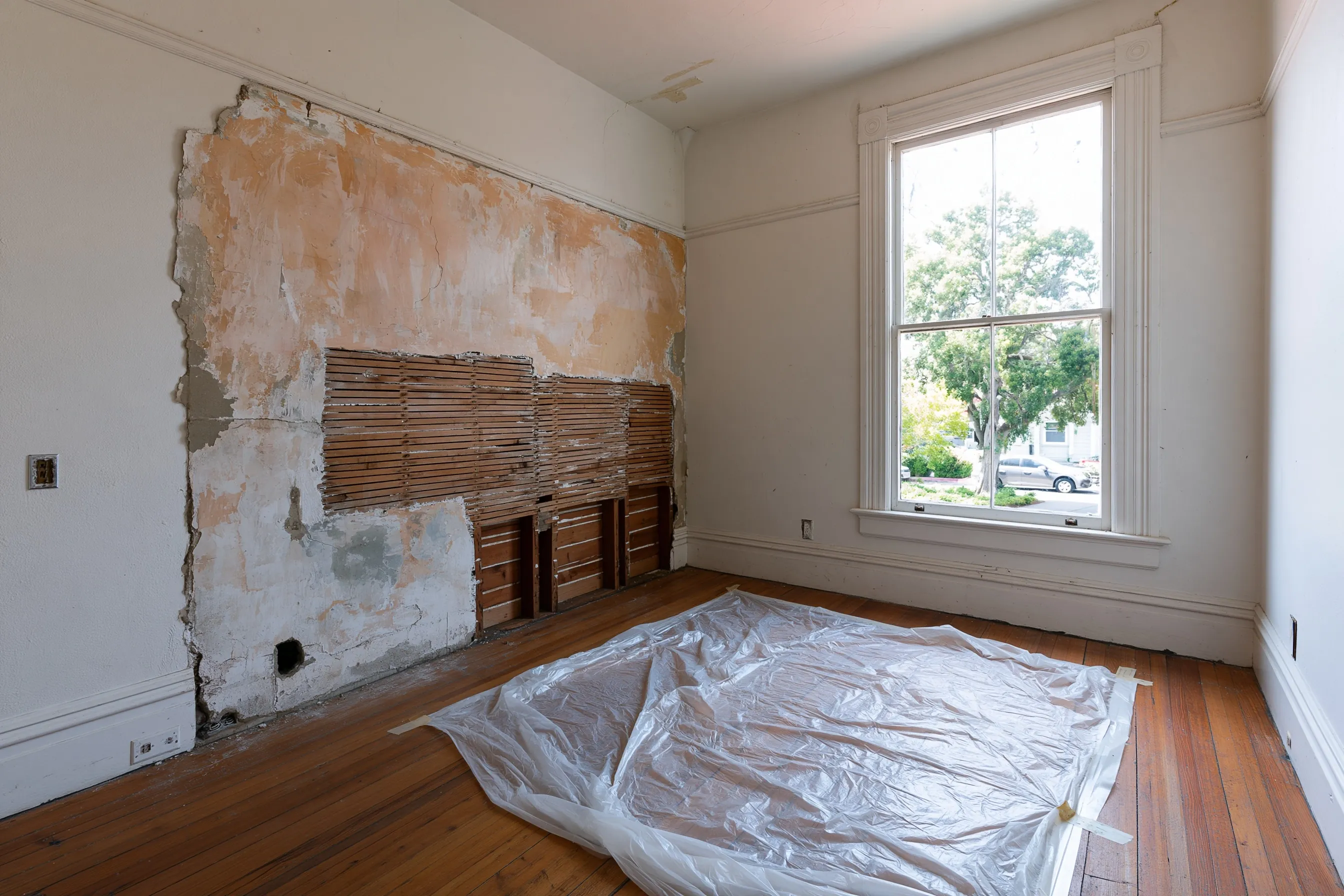 Bedroom with a tall double-hung window, plaster cut away from one wall to show the wood lath, and plastic sheeting over the plank floor